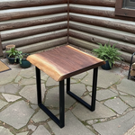 Solid wood live edge side table in walnut color with black steel legs placed on outdoor stone floor near log wall